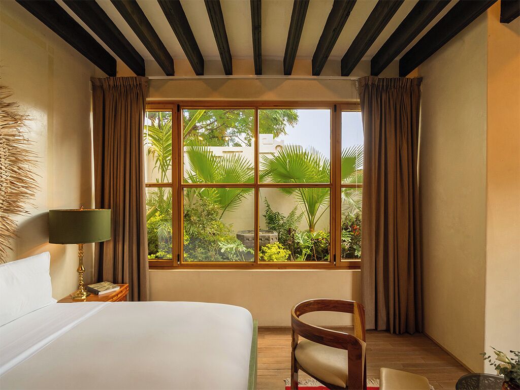 Habitación de hotel con una ventana grande, una cama, una silla y una mesita de noche con lámpara.