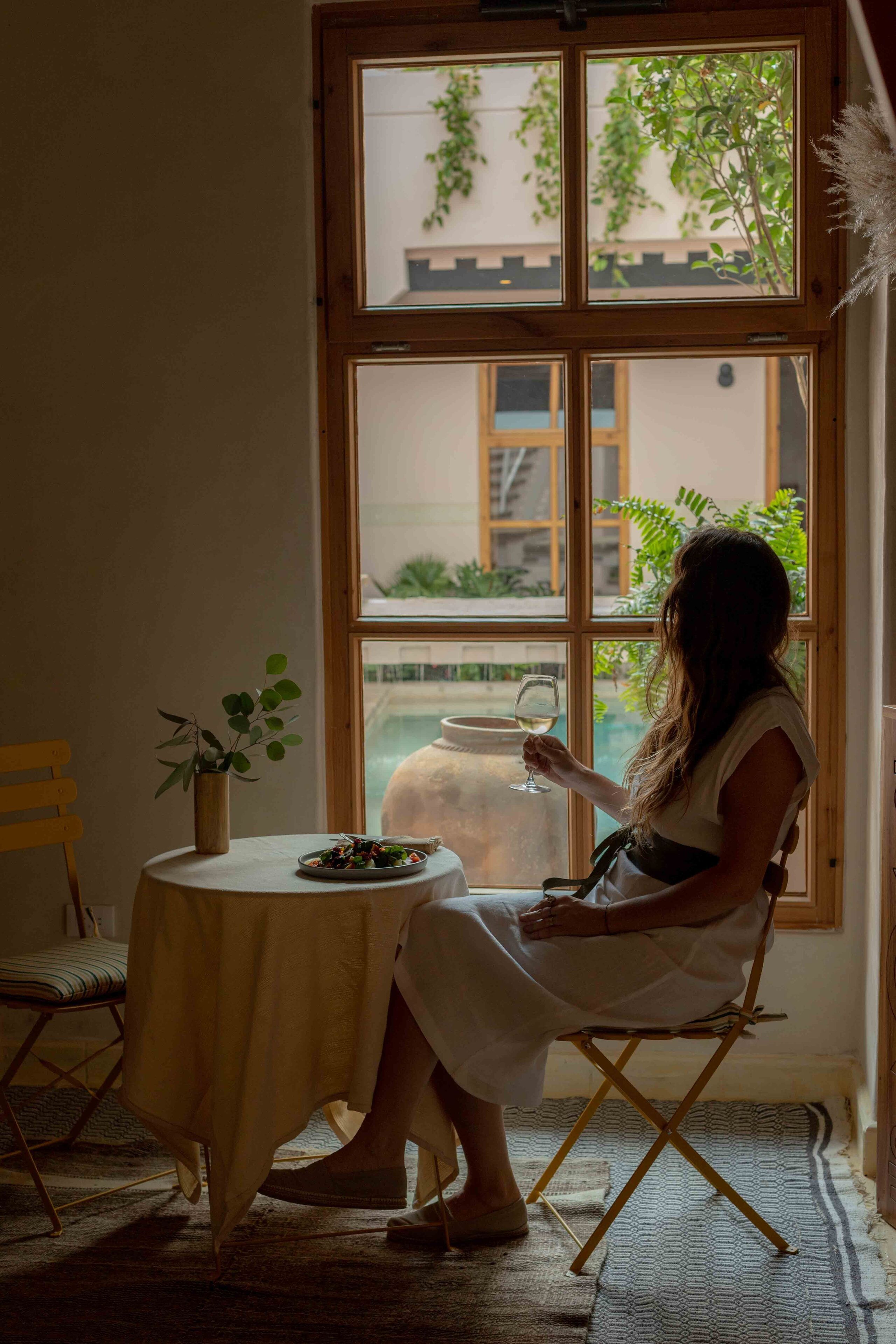 Una mujer sentada a una mesa junto a un ventanal con vistas a un jardín y un edificio.