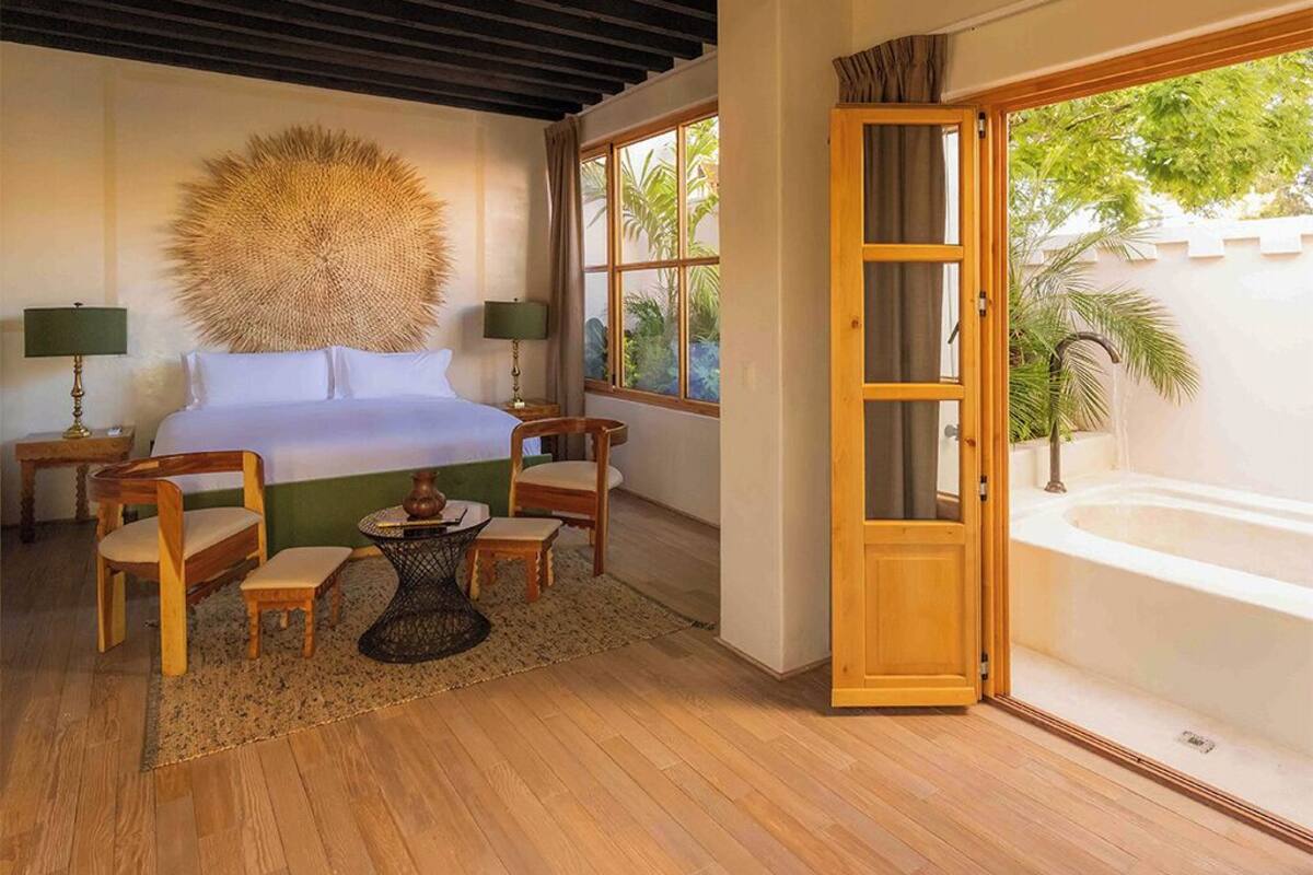 Un dormitorio con cama, dos sillas, una mesita, un elemento decorativo grande en la pared y vista a una piscina a través de un ventanal.