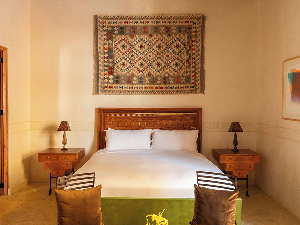 A hotel room with a large bed, wooden headboard, two nightstands, and a colorful tapestry on the wall.