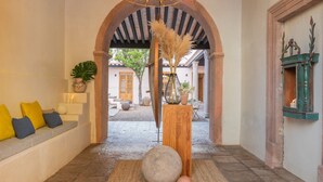 Un pasillo interior con un banco de madera, una araña y un arco rústico que conduce a un espacio exterior.