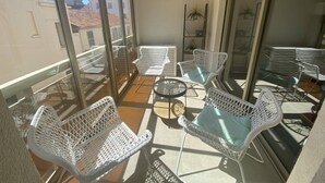 Un balcón con mobiliario de mimbre blanco, incluyendo sillas y una mesa, y amplias puertas de vidrio que dan a una vista urbana.