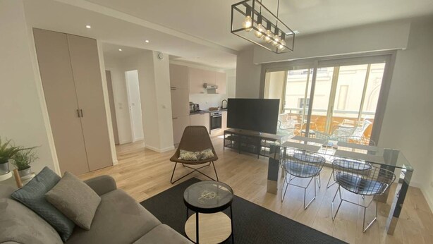 Una sala de estar moderna con un sofá, una televisión, una zona de comedor con sillas y una cocina al fondo.