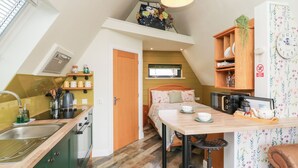 Eine kleine Küche mit Spüle, Herd und Regalen. Angrenzend an die Küche befindet sich ein Essbereich mit Tisch und Stühlen.