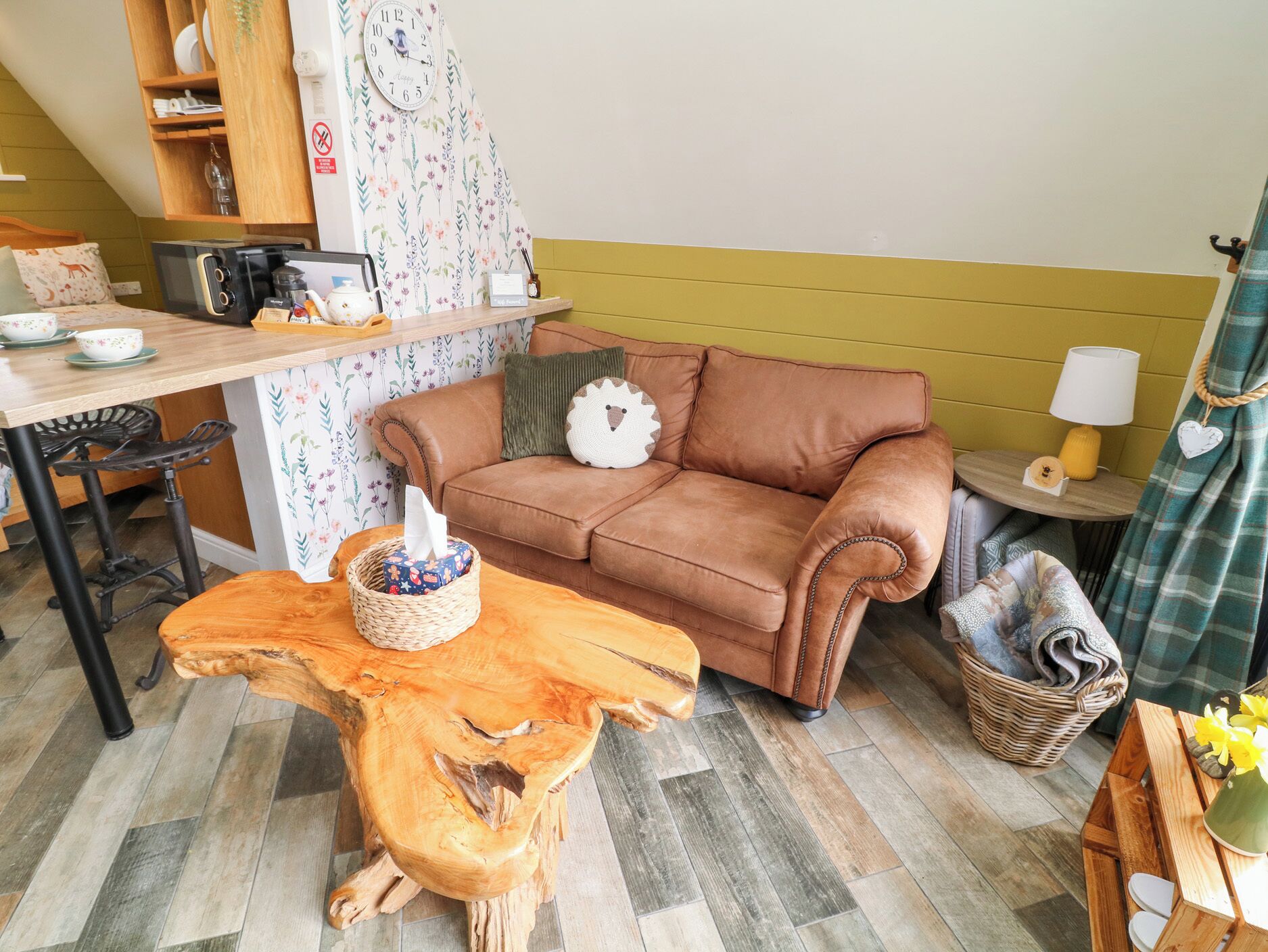 Una sala acogedora con un sofá de cuero marrón, una mesa de centro de madera, una cesta de mimbre y una pequeña mesa redonda con una lámpara.