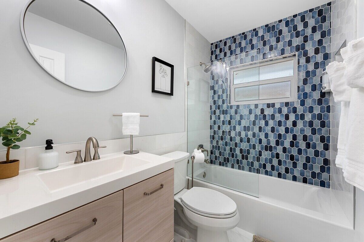Een moderne badkamer met een witte wastafel, een ronde spiegel, een toilet en een douche met blauwe mozaïektegels.