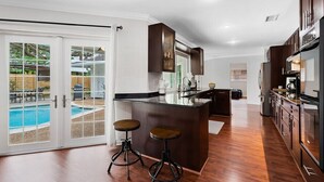 A modern kitchen with a central island, dark wooden cabinets, and a glass backsplash. There is a sliding glass door leading to a pool area. - 15 Mins to Beach! Safe & Quiet Home Near Beach & Downtown~Heated Pool~Ping Pong/Foosball. (Fort Lauderdale)