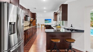 A modern kitchen with a stainless steel refrigerator, wooden cabinets, a central island with stools, and a view of a pool through the window. - 15 Mins to Beach! Safe & Quiet Home Near Beach & Downtown~Heated Pool~Ping Pong/Foosball. (Fort Lauderdale)
