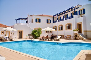 A swimming pool with lounge chairs and umbrellas, surrounded by a building with blue balconies and white walls. - Venardos Hotel & Spa (Kithira)