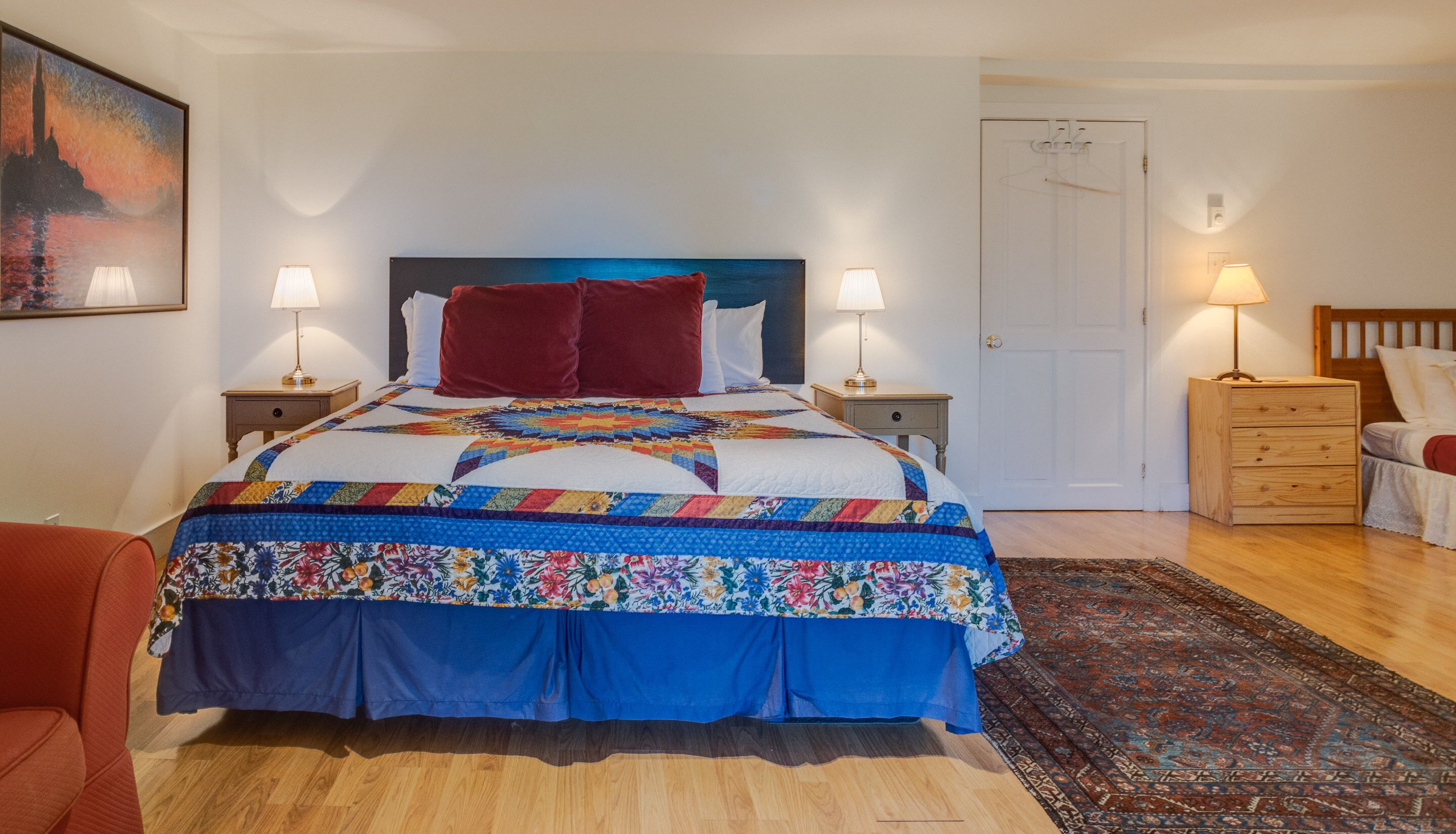 Een net opgemaakt bed met een kleurrijke quilt, twee bedlampjes, een houten nachtkastje, een rode stoel en een deur met een jas er aan.