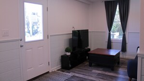 A room with a white door, a black entertainment unit with a TV, a wooden coffee table, and a window with dark curtains. - Beautiful new property, great location. (Waves)