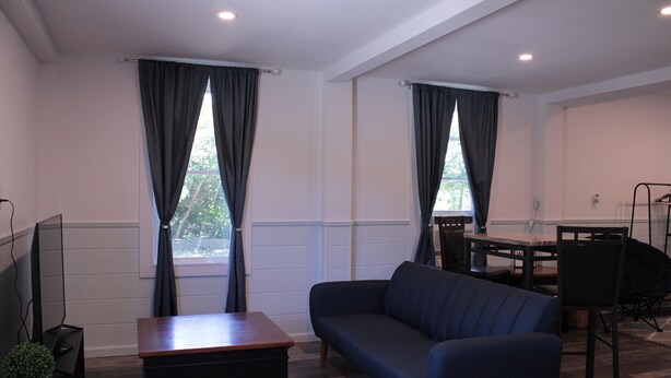 A modern living room with a sofa, a small table, and a TV. There are two windows with curtains, a dining table with chairs, and a small plant on a side table. - Beautiful new property, great location. (Waves)