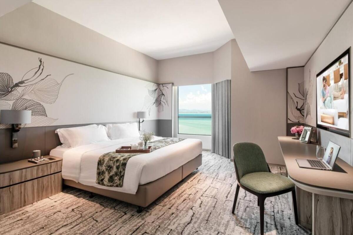 A modern hotel room with a large bed, a desk with a laptop, a chair, and a view of the sea.