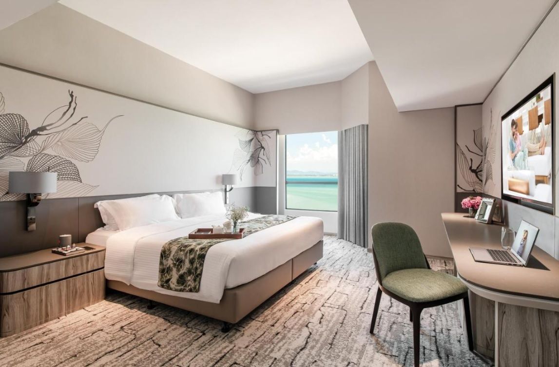 A modern hotel room with a large bed, a desk with a laptop, a chair, and a view of the sea.