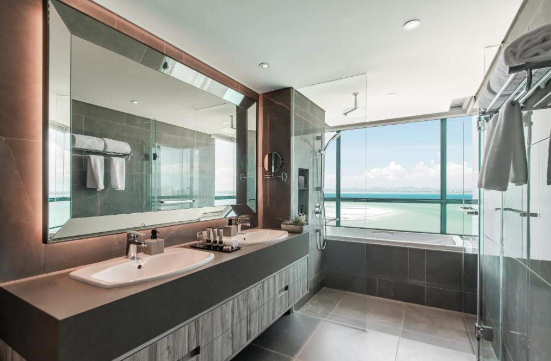 A modern bathroom with a large mirror, two sinks, a shower, and a view of the sea.