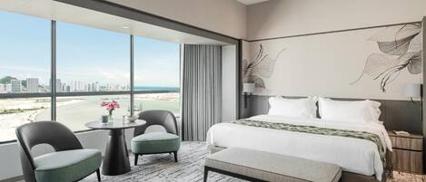 A hotel room with a large window, a bed, a round table, two chairs, and a bench.