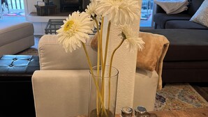 En stue med en glaskrukke med hvide blomster på et træbord, en sofa og en pejs i baggrunden.