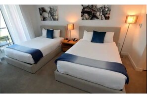 Two single beds with matching headboards and footboards, adorned with white linens and throw pillows, sit beside bedside tables with lamps.