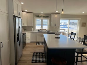 Eine moderne Küche mit weißen Schränken, einem Edelstahlkühlschrank, einer großen Kochinsel und Blick auf den Strand durch die Fenster.