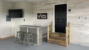 Una habitación con pared de madera, puerta negra, letrero de bienvenida, televisor montado en la pared y una zona de bar con taburetes.