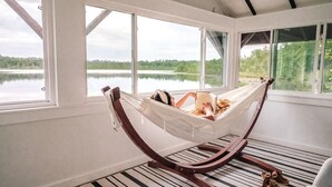 Eine Hängematte in einem Zimmer mit großen Fenstern und Blick auf einen See, Holzboden und eine Aussicht ins Freie.