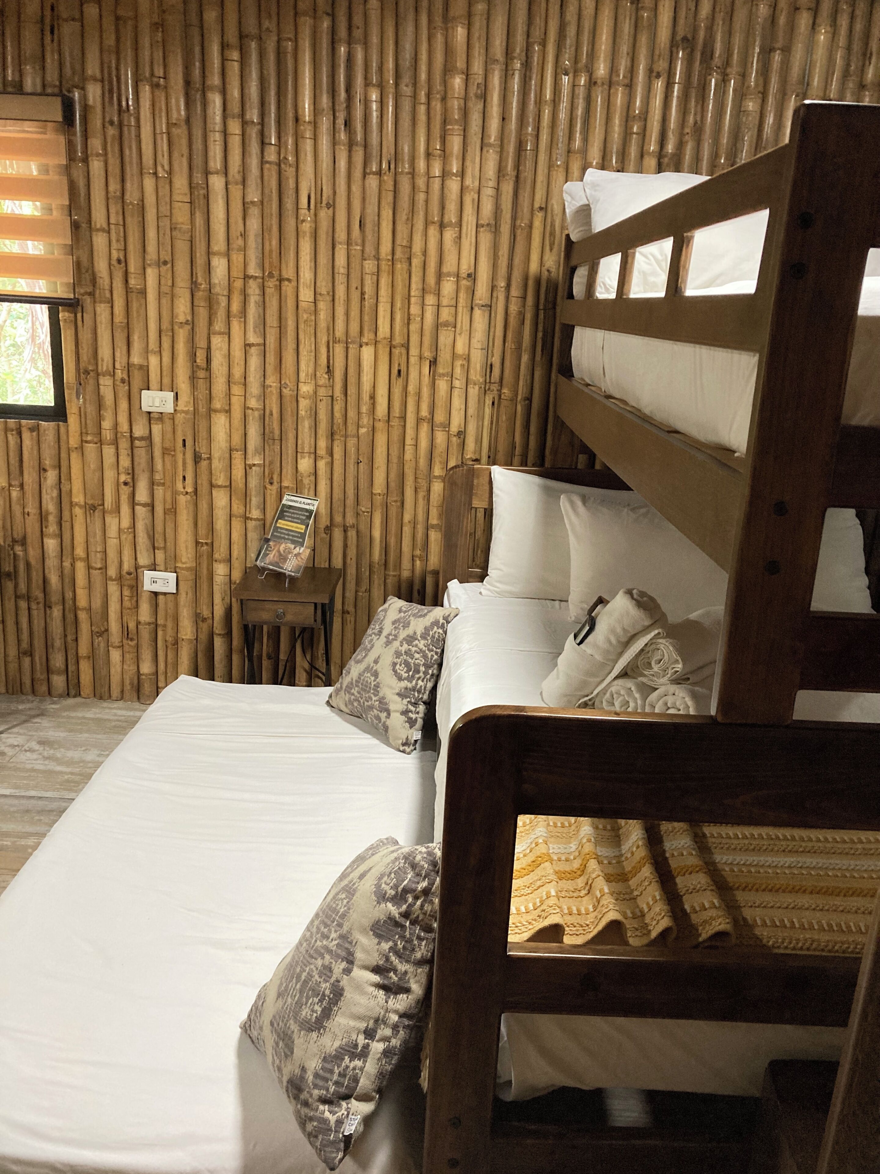 A room with a bed, pillows, a small table with a sign, and a bamboo wall.