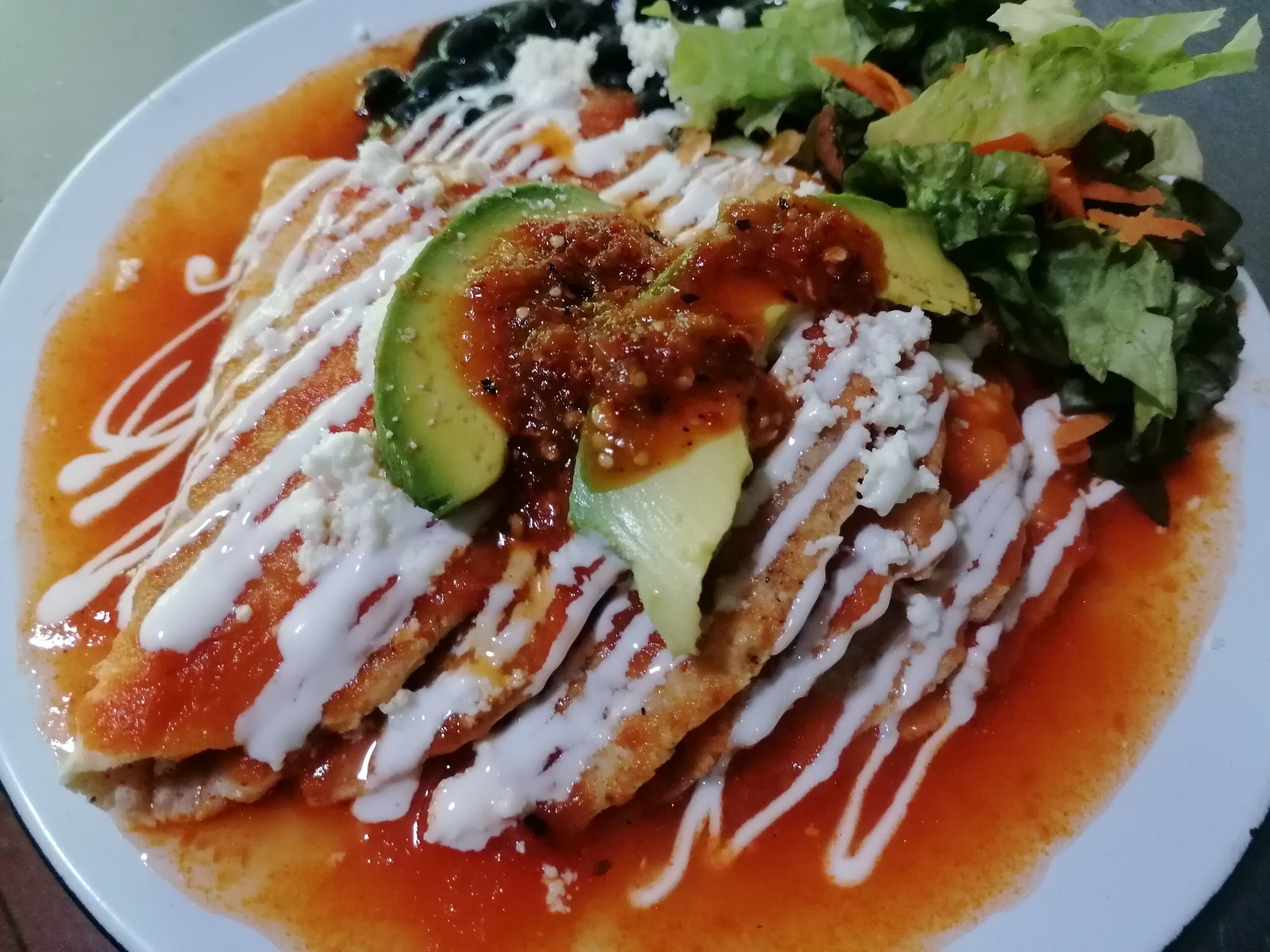 A plate of enchiladas topped with avocado, sour cream, and a red sauce, accompanied by a side salad.