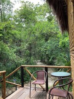 Una terraza de madera con dos sillas de mimbre y una mesa redonda pequeña, con vista a un bosque frondoso.