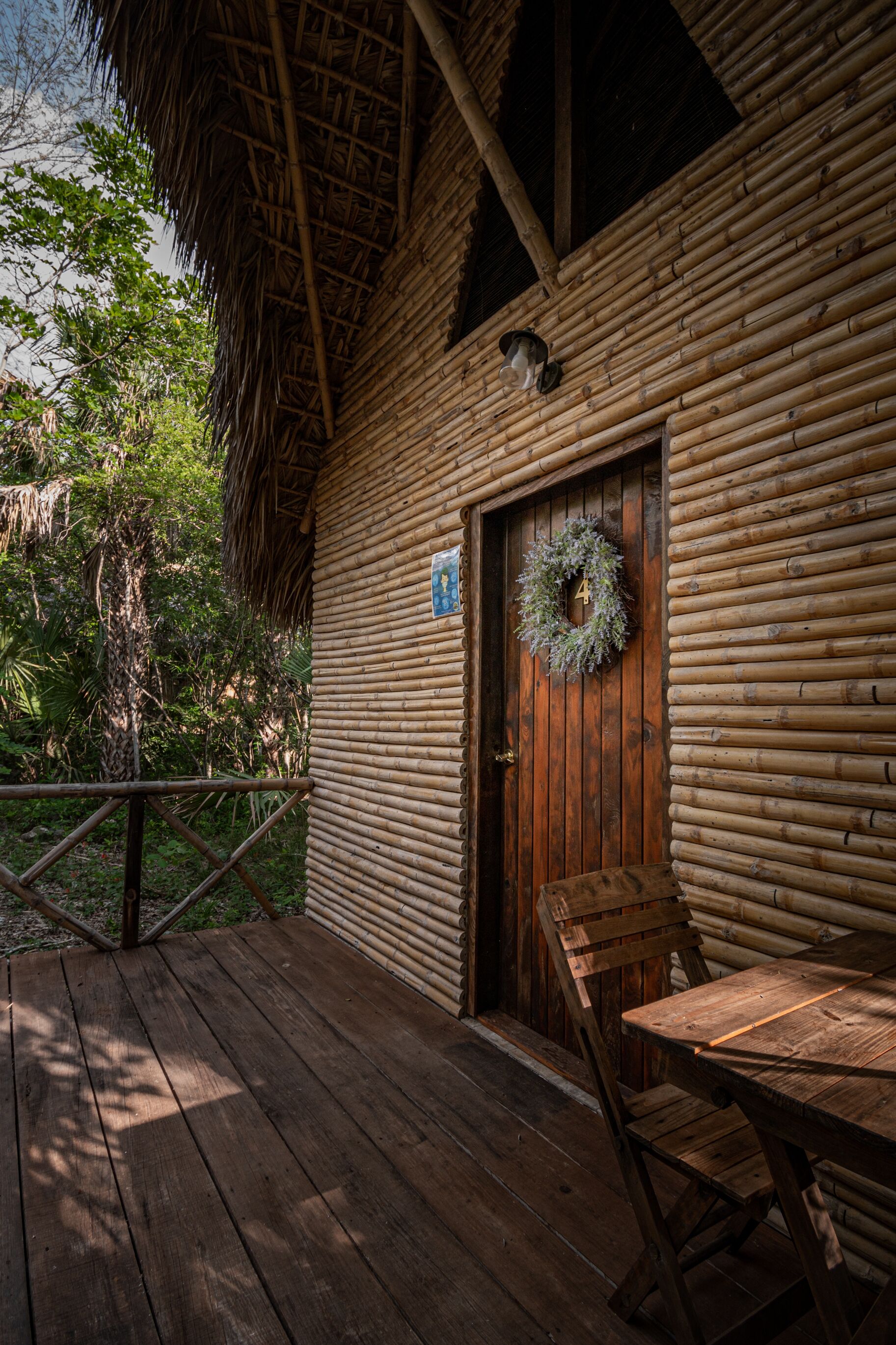 A rustic wooden cabin with a thatched roof, a wooden door with a wreath, and a wooden deck with a table and chairs.