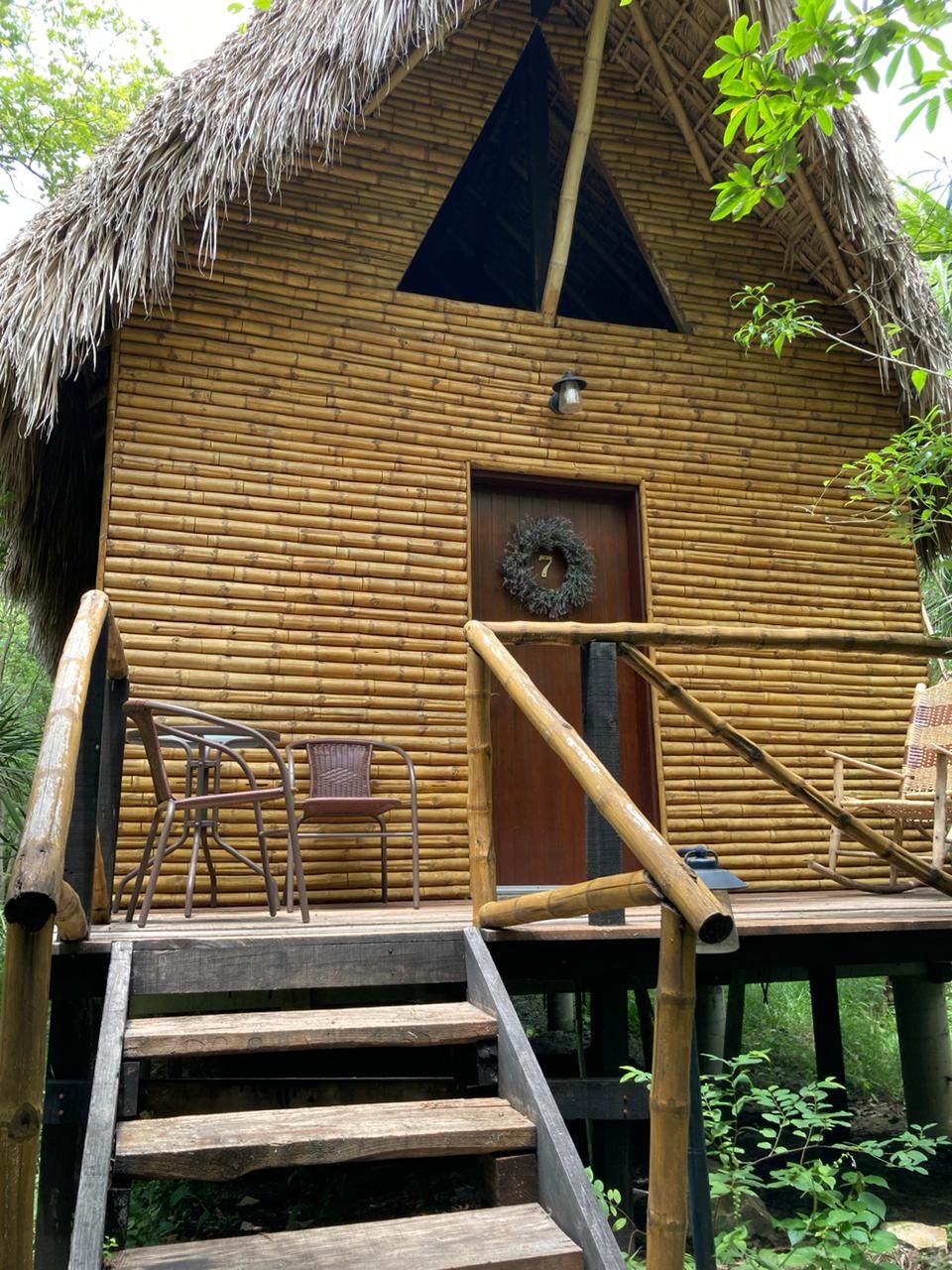 A rustic wooden cabin with a thatched roof, a wooden door with a wreath, and a set of stairs leading to a deck with chairs.