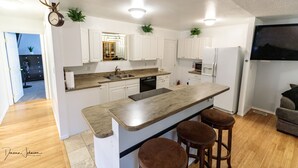 Una cocina moderna con armarios blancos, encimera de granito y lavavajillas integrado. Hay dos taburetes en la isla y un reloj con cuernos en la pared.