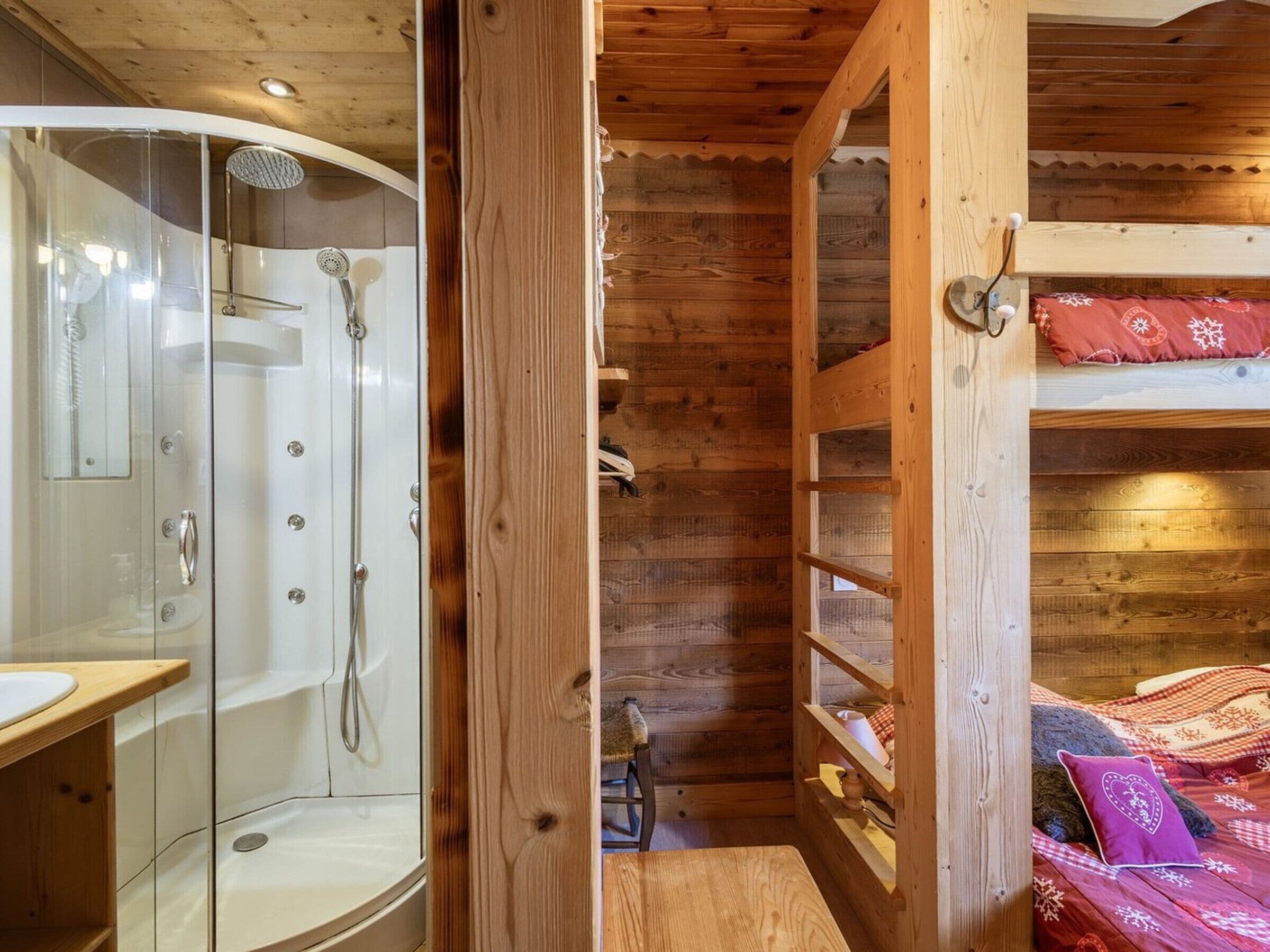 Interior de una cabaña de madera con ducha separada por vidrio, una litera con manta roja estampada y un estante de madera con varios objetos.