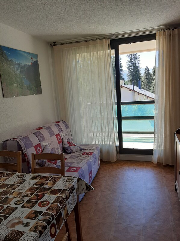 A cozy room with a bed, a sofa, and a table with a coffee-themed tablecloth. There is a painting on the wall and a window with curtains.
