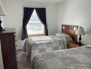 A bedroom with two beds, a wooden dresser, a nightstand with a lamp, and a window with curtains.