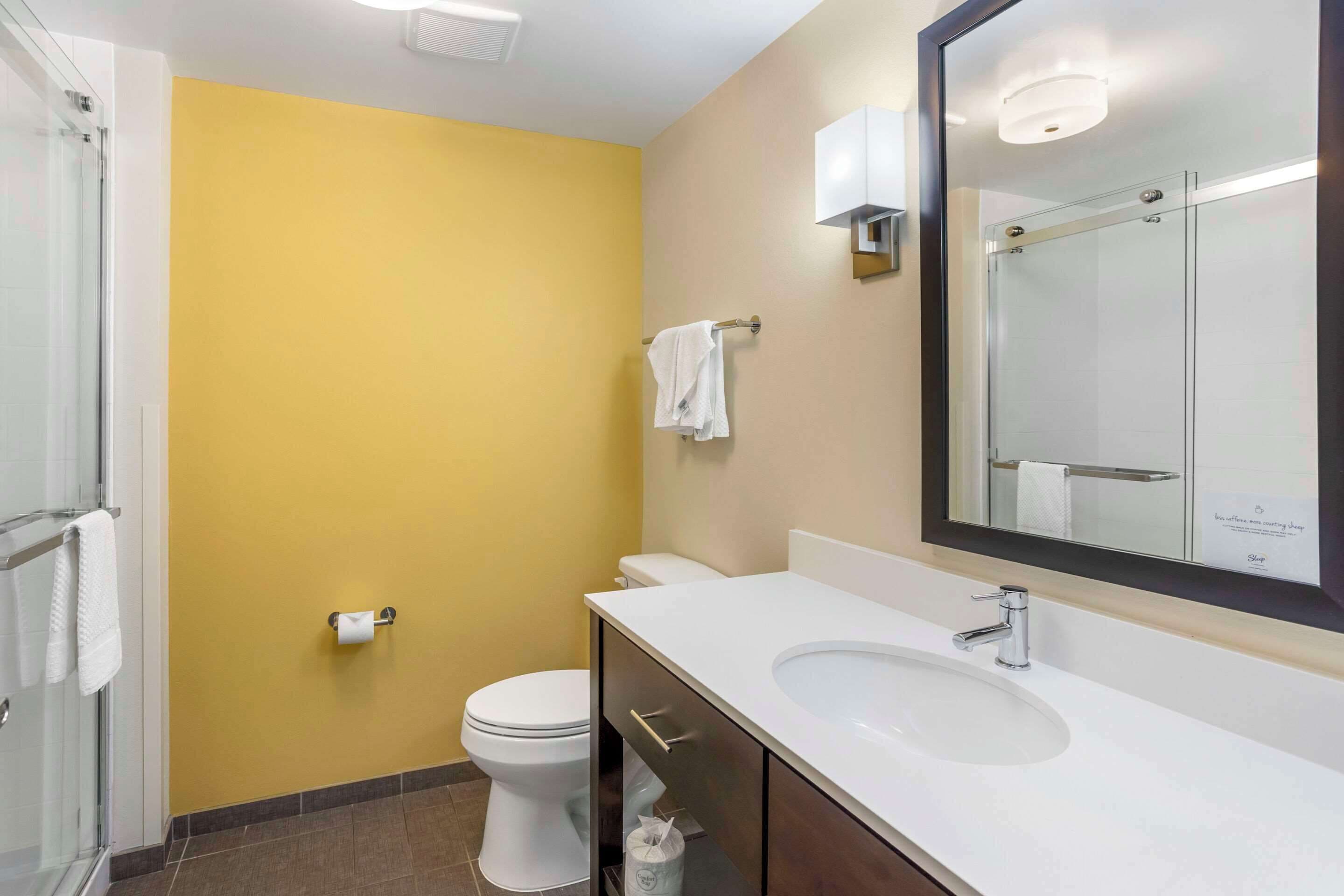 Un baño con pared amarilla, inodoro blanco y un espejo grande sobre el lavabo.