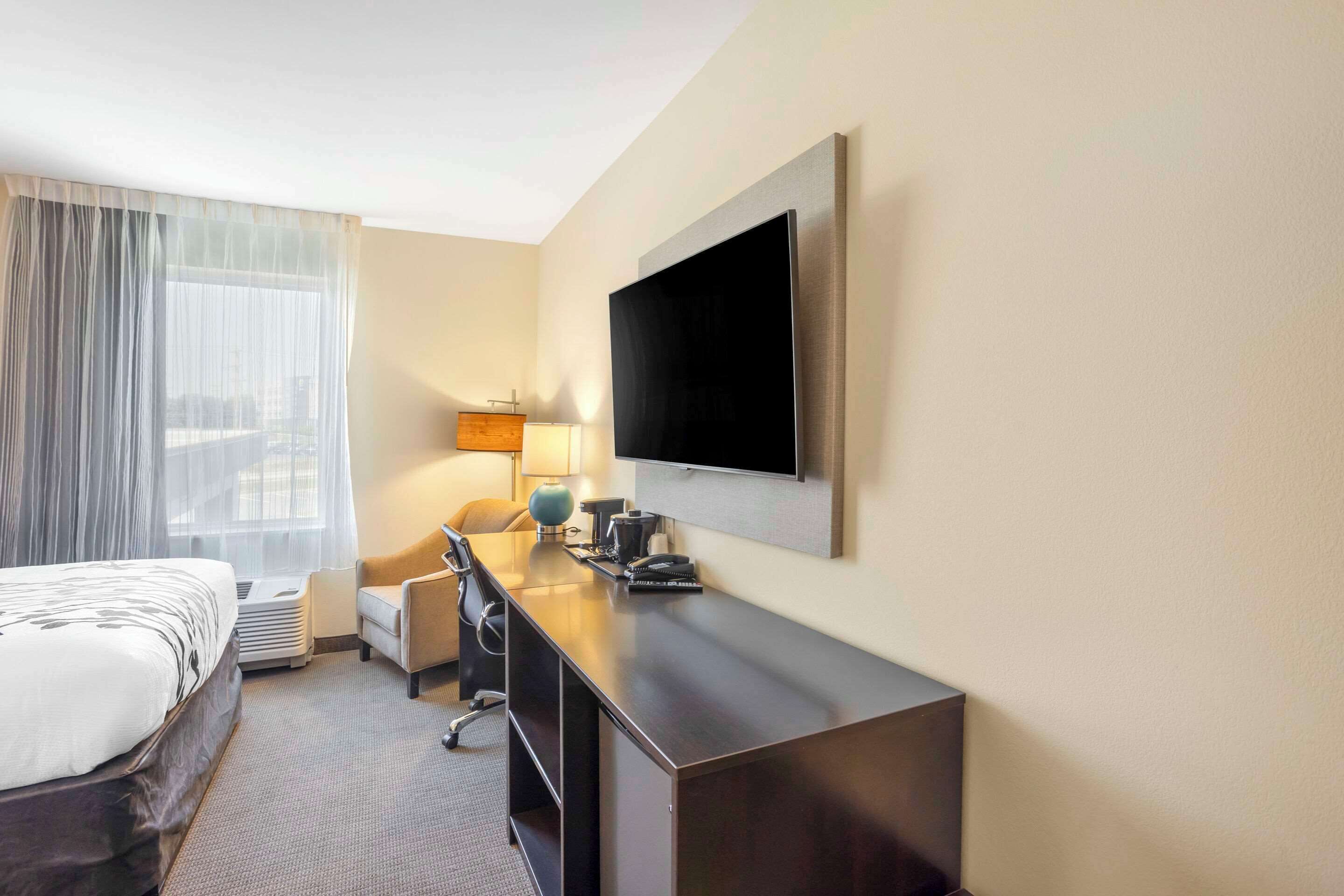 Habitación de hotel con cama, escritorio, silla, lámpara y televisor de pantalla plana montado en la pared.