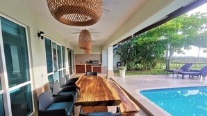 Ein Poolbereich mit Holztisch und -stühlen, einer hängenden Lampe und Blick auf Pool und Garten.