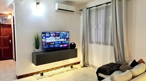 A modern living room with a flat-screen TV mounted on a wall, a sofa, and a window with curtains. - A luxurious 2 bedroom apartment w/ a king size bed (Mbezi Beach)
