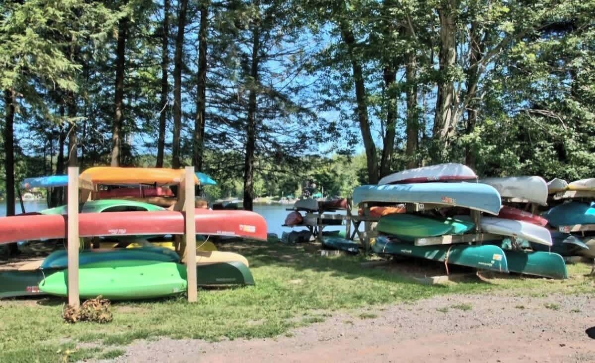 A collection of canoes and kayaks stacked on a grassy area near a body of water, surrounded by trees.
