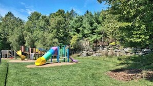 A playground with slides and climbing structures, surrounded by trees and a stone wall.