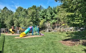 Un parque infantil con toboganes y estructuras para escalar, rodeado de árboles y un muro de piedra.