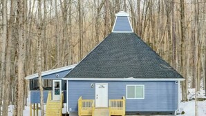 A small, single-story cabin with a dark roof and light blue siding, surrounded by a forest.