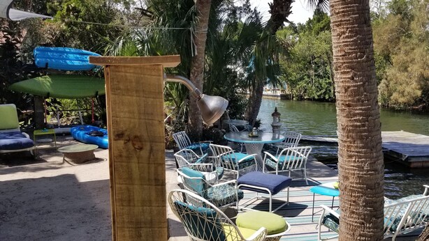 Un espace extérieur aménagé avec des chaises et des tables en métal blanc, entouré de palmiers et bordé par une étendue d’eau.