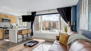 Un apartamento moderno con cocina, comedor y sala de estar. Cuenta con un amplio ventanal con vista a la ciudad, un televisor de pantalla plana y un rincón de estar acogedor con un sofá de cuero y una mesa de centro.