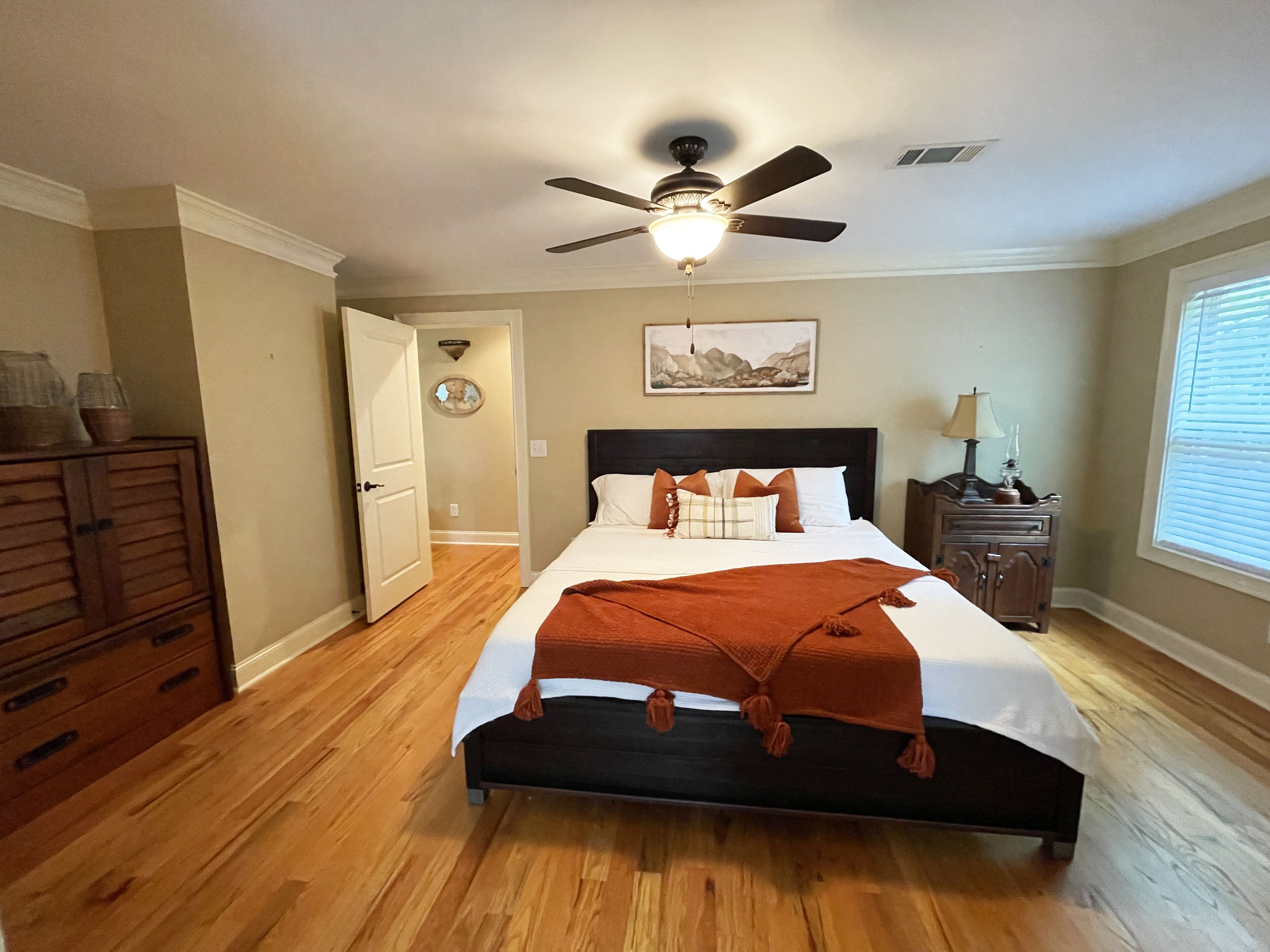 A bedroom with a bed, a ceiling fan, a nightstand, a dresser, a window with blinds, and a door leading to another room.