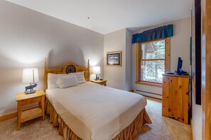 A bedroom with a large bed, two bedside tables with lamps, a wooden headboard, a window with a view of trees, and a wooden dresser. - Charming Condo Nestled in the East Keystone Resort Neighborhood, Shuttle to Ski Slopes, Outdoor Pool and Hot Tub (Keystone)