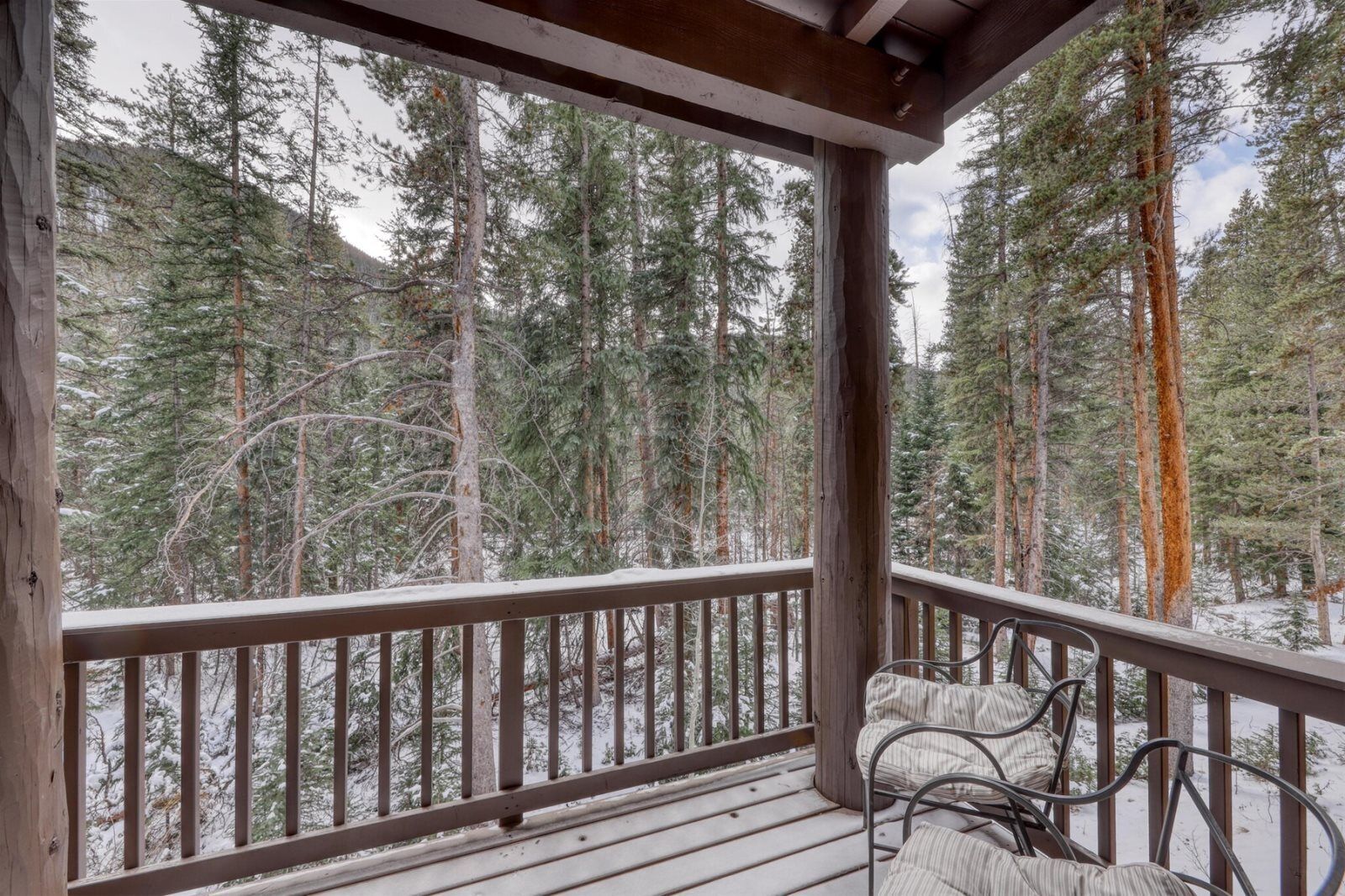 A wooden deck with metal chairs, overlooking a snowy forest.