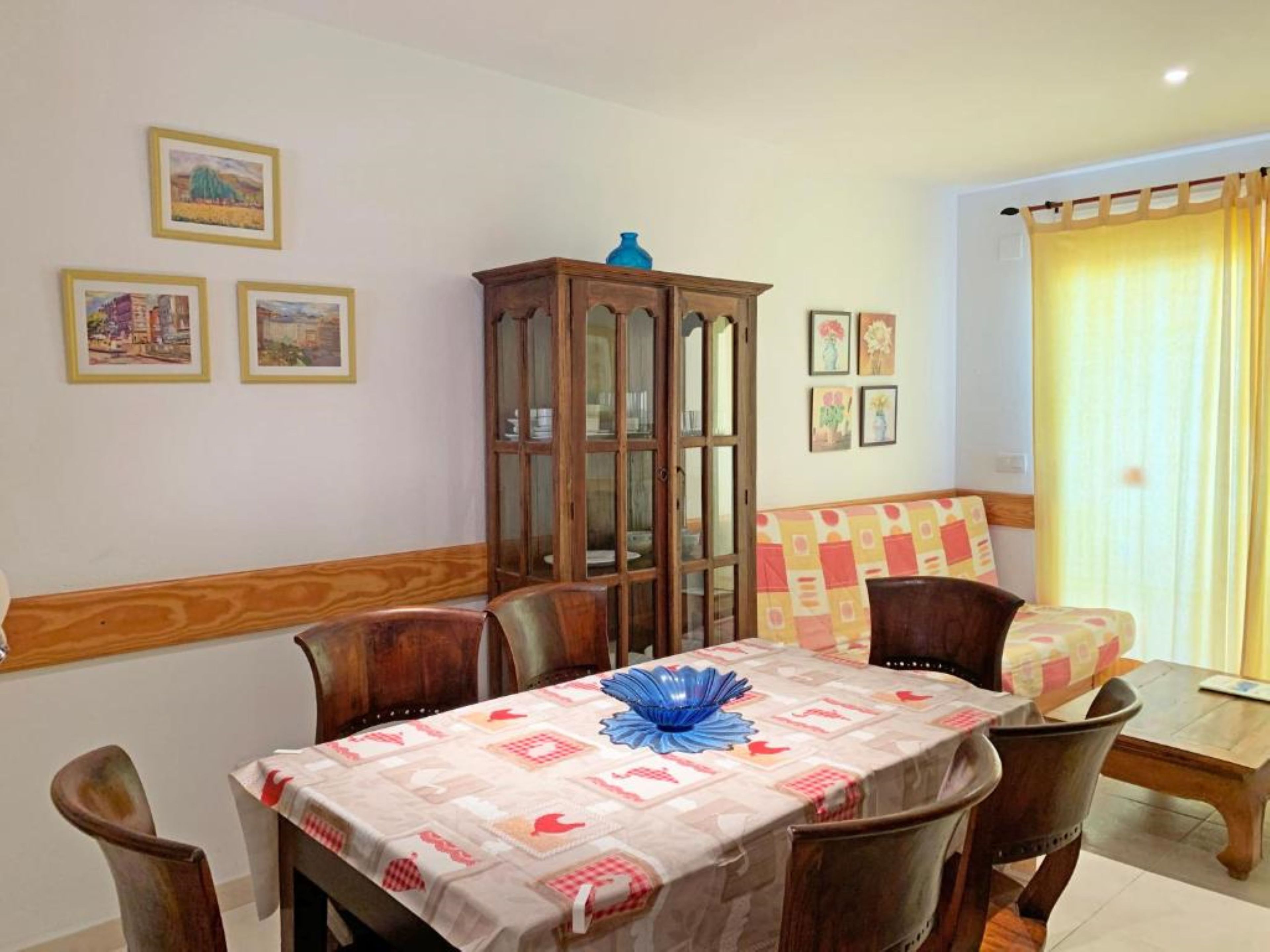 A dining area with a table set for four, surrounded by wooden chairs. There is a cabinet with glass doors against the wall, and a window with curtains.