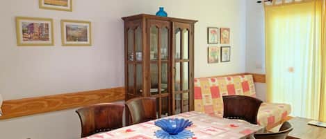 A dining area with a table set for four, surrounded by wooden chairs. There is a cabinet with glass doors against the wall, and a window with curtains.