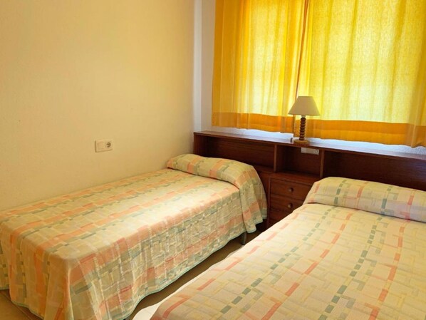 A small, simple room with two single beds, a nightstand, a lamp, and a window with yellow curtains.